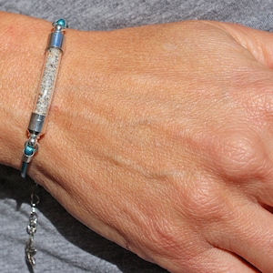 Pode incluir: Uma pulseira de prata com um tubo de vidro transparente preenchido com areia. A pulseira tem detalhes azuis e uma extens&atilde;o de corrente. A areia parece ser uma mistura de gr&atilde;os claros e escuros. A pulseira &eacute; usada no pulso.