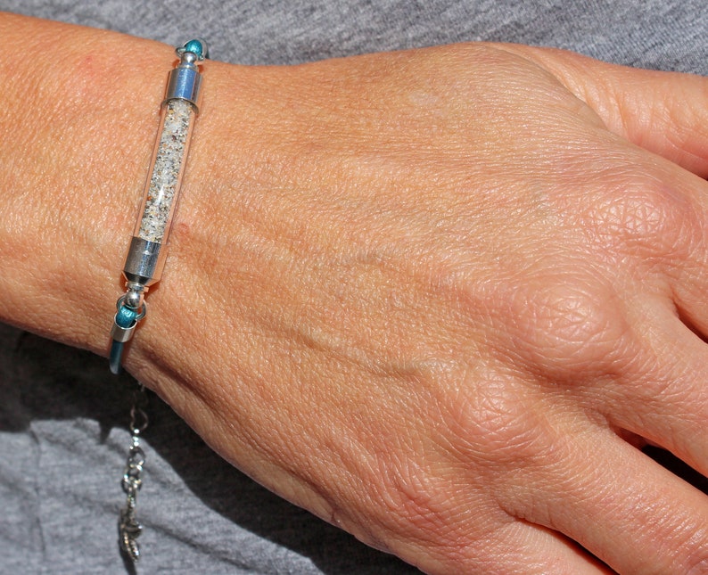 Pode incluir: Uma pulseira de prata com um tubo de vidro transparente preenchido com areia. A pulseira tem detalhes azuis e uma extens&atilde;o de corrente. A areia parece ser uma mistura de gr&atilde;os claros e escuros. A pulseira &eacute; usada no pulso.