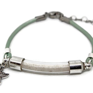 Pode incluir: Uma pulseira com um tubo transparente preenchido com areia, conectada por detalhes met&aacute;licos prateados e um cord&atilde;o verde claro. Um pingente de estrela do mar pende da pulseira, que tem uma corrente ajust&aacute;vel.