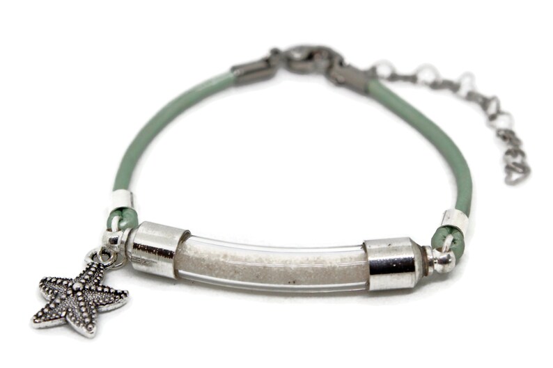 Pode incluir: Uma pulseira com um tubo transparente preenchido com areia, conectada por detalhes met&aacute;licos prateados e um cord&atilde;o verde claro. Um pingente de estrela do mar pende da pulseira, que tem uma corrente ajust&aacute;vel.