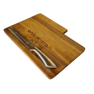 May include: A wooden cutting board with a bread knife. The board has a rectangular shape with a smaller, attached section. The knife has a serrated blade and a silver handle. Hebrew text is engraved on the board.