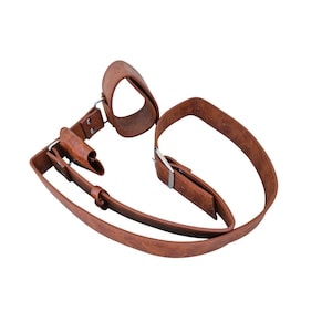 Puede incluir: Una correa de cuero marrón con una muñequera, una funda central y una correa con una hebilla plateada. El cuero es de un color marrón rojizo cálido. El diseño sugiere un artículo funcional, posiblemente vintage.