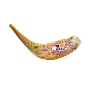 May include: A curved shofar, a traditional Jewish instrument, with a painted lion's face and the words "Lion of Judah." The shofar is yellow with brown and orange hues, and the lion's face has red, white, and black details.