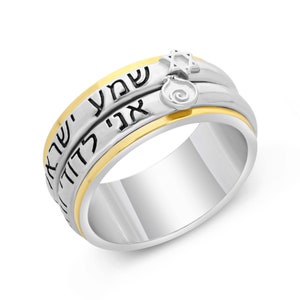 Puede incluir: Anillo plateado y dorado con texto hebreo. El anillo presenta una Estrella de David y un dije de granada. El diseño incluye una banda dorada entre dos bandas plateadas, con letras hebreas grabadas. Una joya única.