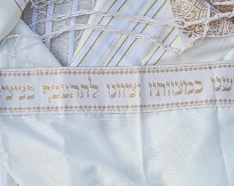 Puede incluir: Talit blanco con rayas doradas y un borde bordado en oro. El borde presenta el texto hebreo "Bendito seas, Señor nuestro Dios, Rey del universo."