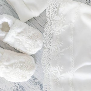 Pu&ograve; includere: Scarpine da beb&egrave; bianche con bordo in pizzo e una coperta bianca con bordo in pizzo. Le scarpine e la coperta sono su una superficie di legno bianca.