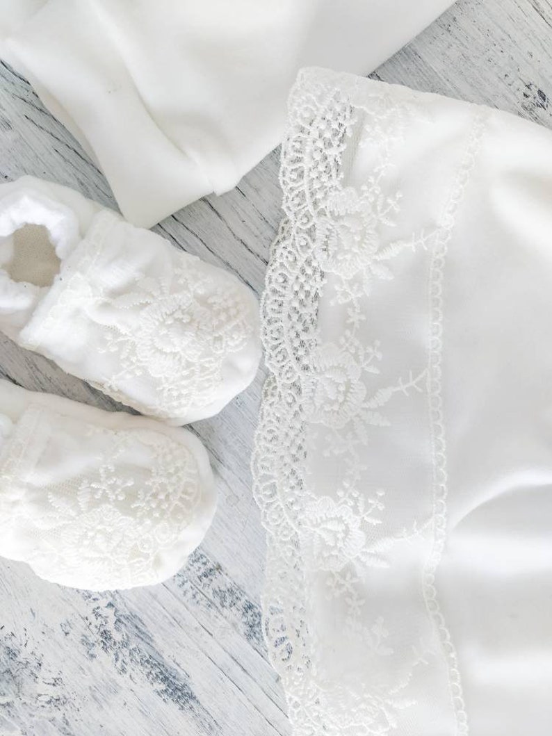 Pu&ograve; includere: Scarpine da beb&egrave; bianche con bordo in pizzo e una coperta bianca con bordo in pizzo. Le scarpine e la coperta sono su una superficie di legno bianca.