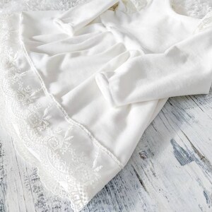 Pu&ograve; includere: Abito bianco per neonata con bordo in pizzo. L'abito ha maniche lunghe e una gonna a pieghe.