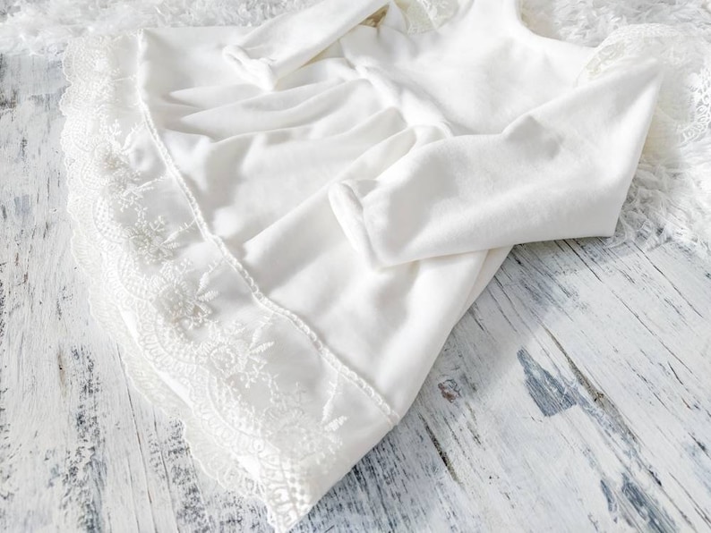 Pu&ograve; includere: Abito bianco per neonata con bordo in pizzo. L'abito ha maniche lunghe e una gonna a pieghe.