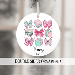 May include: A white round Christmas ornament with a pink and blue book stack design and pink bows. The ornament has the name "Tracy" and the year "2024" printed on it. The ornament is labeled as a double-sided ornament.