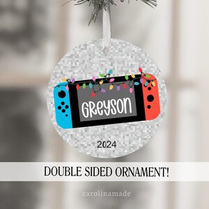 May include: A round, silver glitter Christmas ornament with a cartoon image of a Nintendo Switch console with colorful lights. The console screen says "Greyson" and the year "2024" is printed below the console. The ornament is double-sided.
