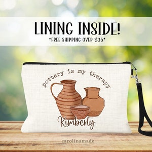 May include: White canvas zippered pouch with a black zipper and a black strap. The pouch has a brown and white illustration of pottery with the text "pottery is my therapy" and the name "Kimberly" in brown text. The text "carolinamade" is printed in small brown text at the bottom of the pouch.