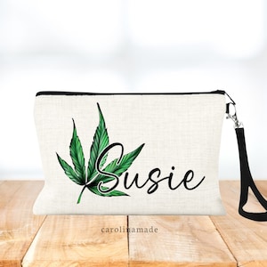 May include: A white canvas makeup bag with a black zipper and a black strap. The bag has a green marijuana leaf design and the name "Susie" written in black cursive. The bag is on a wooden surface.