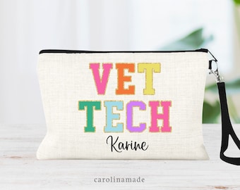 May include: A white canvas makeup bag with a black zipper and a black strap. The bag has the words "VET TECH" in colorful letters with a gold outline. The name "Karine" is written in gold below the words. The words "carolinamade" are printed in black at the bottom of the bag.