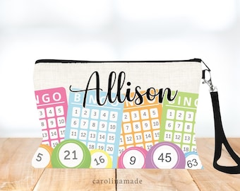 May include: A beige canvas zippered pouch with a black strap. The pouch has a colorful bingo card design with the name "Allison" printed in black cursive. The pouch has several colorful bingo balls with numbers printed on them.