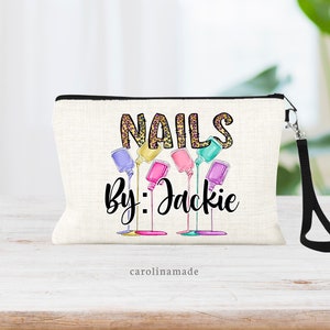 May include: White canvas makeup bag with a black zipper and a black strap. The bag has a design of colorful nail polish bottles dripping with the text "NAILS by: Jackie" in black and gold lettering. The text "carolinmade" is printed in black at the bottom of the bag.