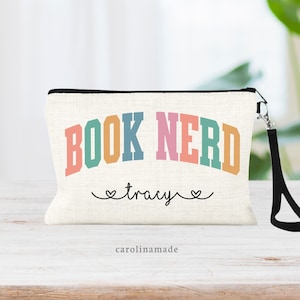 May include: A white canvas makeup bag with a black zipper and a black strap. The bag has the words "BOOK NERD" printed in rainbow colors in an arched font. The words "tracy" are printed in black cursive below the rainbow text. The words "carolinmade" are printed in black below the cursive text.