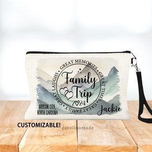 May include: A white canvas zippered pouch with a black zipper and a black strap. The pouch has a watercolor design of mountains and a circle with the text "Family Trip 2024" and "Great Memories, Great Laughs, Great Times, Great Family". The text "Jackie" is below the circle.