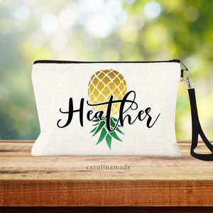 May include: White zippered pouch with a black zipper and a black strap. The pouch has a gold and green pineapple design with the name "Heather" written in black cursive. The pouch has the text "carolinmade" printed on the bottom.
