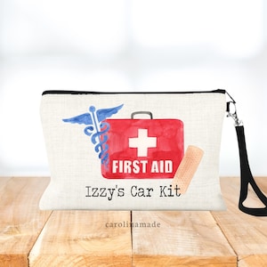 May include: A white canvas zippered pouch with a black zipper and a black strap. The pouch has a red first aid kit with a white cross and the words "First Aid" printed on it. There is a blue medical symbol and a bandage on the pouch. The text "Izzy's Car Kit" is printed on the pouch.