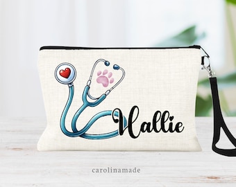 May include: A white canvas makeup bag with a black zipper and a black strap. The bag has a cartoon image of a stethoscope with a red heart and a pink paw print. The text "Hallie" is written in black cursive font. The text "carolinmade" is printed in pink at the bottom of the bag.