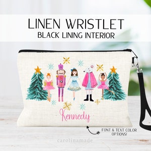 May include: Linen wristlet with a black lining interior. The wristlet features a watercolor design of a Nutcracker, a mouse, and ballerinas in front of Christmas trees. The wristlet is personalized with the name "Kennedy".