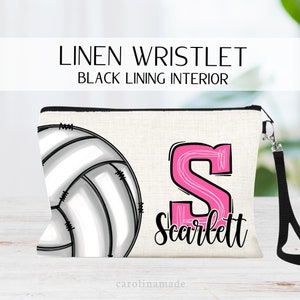 May include: A white linen wristlet with a black lining. The wristlet features a black and white volleyball graphic and a pink letter "S" with the name "Scarlett" written below. The wristlet has a black zipper and a black wrist strap.