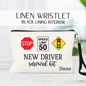 May include: A white linen wristlet with a black lining. It has a black zipper and a black strap. The front of the wristlet has three traffic signs: a stop sign, a speed limit sign that says "50", and a traffic light. The text "NEW DRIVER survival kit" and the name "Dana" are printed on the wristlet.