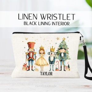 May include: A white linen wristlet with a black lining interior. The wristlet features a watercolor illustration of a Nutcracker, a ballerina, a mouse king, and a Christmas tree. The wristlet is personalized with the name "TAYLOR".