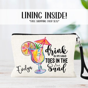 May include: A white canvas makeup bag with a black zipper and a black strap. The bag features a colorful illustration of a cocktail with a lime wedge and an umbrella. The text "drink in my hand toes in the sand" is printed in black. The name "Evelyn" is printed in black below the illustration.
