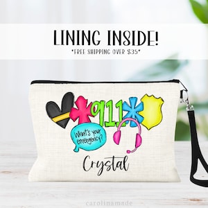 May include: A white canvas makeup bag with a black zipper and a black strap. The bag has a colorful design with a heart, a star, a cross, a police badge, a headset, and the number 911. The text "What's your emergency?" is in a speech bubble. The name "Crystal" is written below the design.