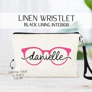 May include: Linen wristlet with black lining, featuring a pink glasses design and the name "Danielle". Includes a black wrist strap and the text "Choose your glasses color". The bag is off-white with a black zipper.