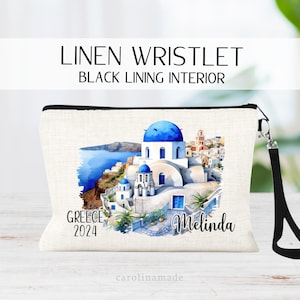 May include: White linen wristlet with a black lining. The wristlet features a watercolor illustration of a white building with a blue dome, surrounded by other white buildings and palm trees. The text "Greece 2024" and "Melinda" is printed on the wristlet.