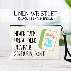May include: A linen wristlet with a black lining interior. The wristlet features a green and blue joker card design with the text "NEVER EVER USE A JOKER IN A PAIR. SERIOUSLY. DON'T."