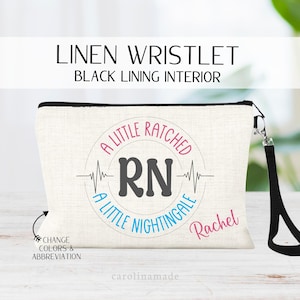 May include: Linen wristlet with black lining. The off-white pouch features the text "A Little Ratched RN A Little Nightingale" in a circular design with a heartbeat line. The name "Rachel" is printed in pink. Includes a black wrist strap.