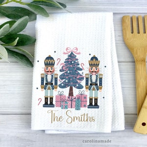 May include: White kitchen towel with a Christmas tree and two nutcracker figures in blue and pink. The text "The Smiths" is printed below the figures.