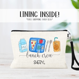 May include: A white canvas lunch bag with a black zipper and a black strap. The bag has a colorful illustration of a lunch tray with a slice of pizza, an apple, peas, a cookie, a carton of milk, a fork, a knife, and a juice box. The text "lunch crew" and the name "SHERYL" are printed on the bag.