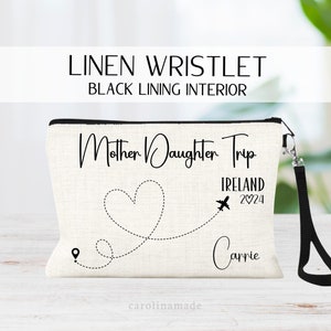 May include: A white linen wristlet with a black lining. The wristlet has a black zipper and a black strap. The front of the wristlet has a black heart with a dotted line leading to a plane. The text "Mother Daughter Trip Ireland 2024 Carrie" is printed on the wristlet.