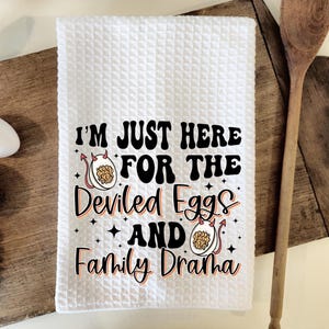 May include: White kitchen towel with a waffle weave texture. The towel has a black and orange graphic with the text "I'm just here for the deviled eggs and family drama".