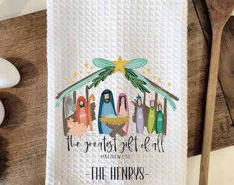 May include: White kitchen towel with a colorful illustration of the nativity scene. The text reads "The greatest gift of all" with a scripture reference and "The Henrys".