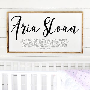 May include: A white wooden sign with a brown frame, featuring the names "Aria Sloan" in black cursive script. Below the names is a black and white Bible verse from Numbers 6:24-26, which reads: "May the Lord bless you and protect you. May the Lord smile on you and be gracious to you. May the Lord show you his favor and give you his peace."