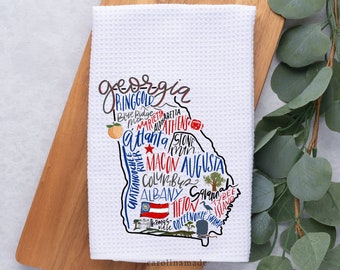 Może przedstawiać: Biały ręcznik kuchenny o strukturze wafelka z kolorową mapą stanu Georgia, USA. Mapa zawiera nazwy miast, takie jak Atlanta, Augusta i Savannah, wraz z ilustracjami brzoskwini, flagi i innych symboli stanowych. Ręcznik jest prezentowany na drewnianej desce.