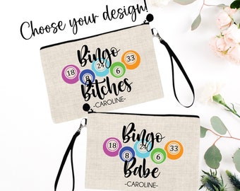 May include: Two beige zippered pouches with black zipper pulls and black straps. The pouches have a linen-like texture and feature a design of colorful bingo balls with numbers and the text "Bingo Bitches -CAROLINE- " and "Bingo Babe -CAROLINE-"