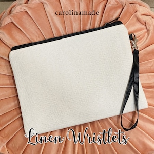 May include: A white linen wristlet with a black zipper and a black wrist strap. The wristlet is on a pink background. The text "Linen Wristlets" is written below the wristlet.