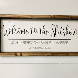 May include: A white wooden sign with a brown frame. The sign says "Welcome to the Shitshow" with the names "John, Rebecca, Samuel, Harper" and "Established 2010" underneath.