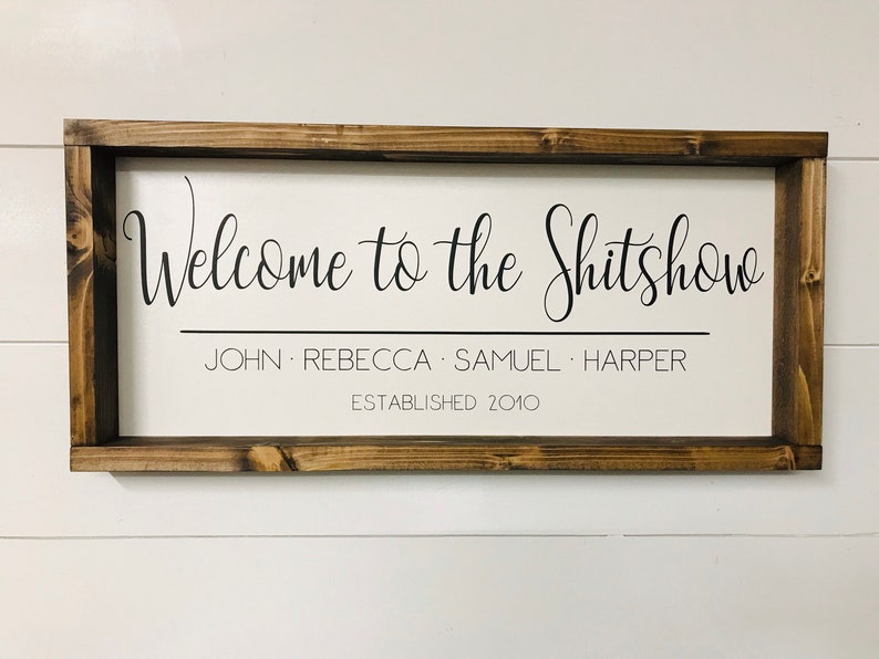 May include: A white wooden sign with a brown frame. The sign says "Welcome to the Shitshow" with the names "John, Rebecca, Samuel, Harper" and "Established 2010" underneath.