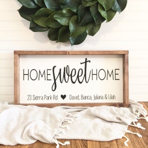 May include: A rectangular wooden sign with the words "HOME sweet HOME" in black script. Below the text is an address and names. The sign is set against a white background and a green wreath.