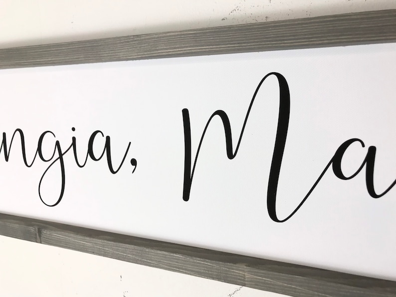 May include: A framed sign with the text "Ingia, Ma" in black cursive script on a white background. The frame is a dark gray wood.
