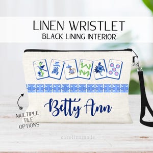 May include: Linen wristlet with black lining, featuring a blue and white Mahjong tile design. The wristlet has a black wrist strap and the name "Betty Ann" in blue script. The text "Multiple Tile Options" is also visible.