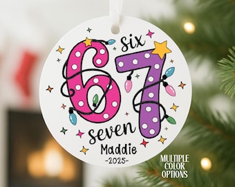 May include: A white, round Christmas ornament with the numbers "6" and "7" in pink and purple, decorated with string lights and stars. The ornament says "six seven" and "Maddie -2025". A ribbon is attached for hanging.
